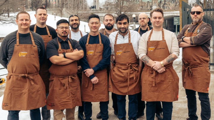 The ten Canadian Culinary Championship 2024 chef contenders stand outside in their tan aprons. Canada's Parliement Hill in Ottawa is visible in the background.
